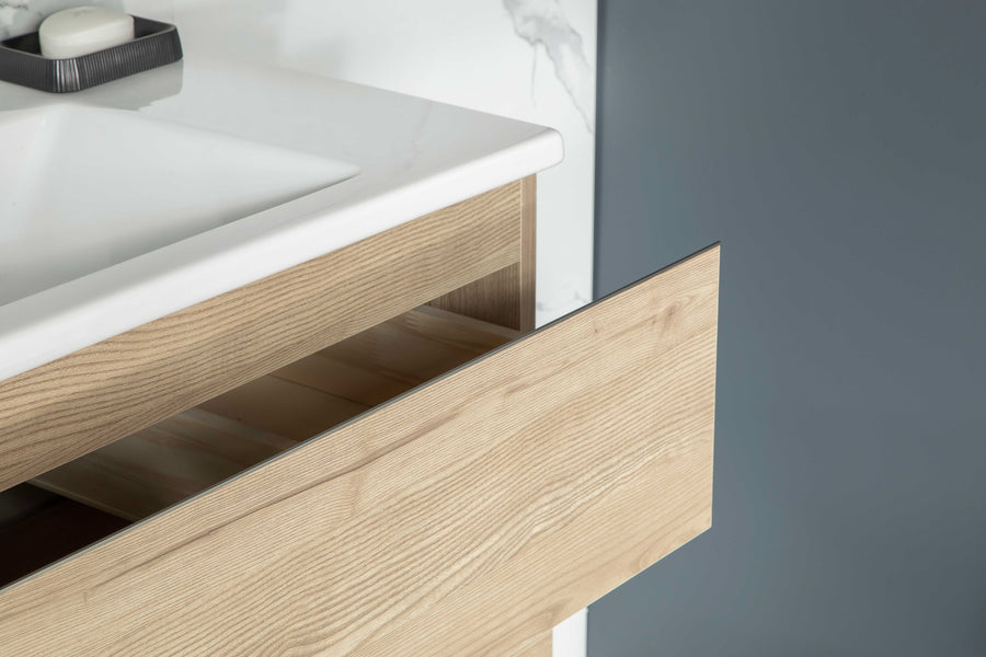 A close-up of a modern bathroom vanity with a white countertop, an open light wood drawer, and a gray wall in the background. A soap dish sits on the counter.