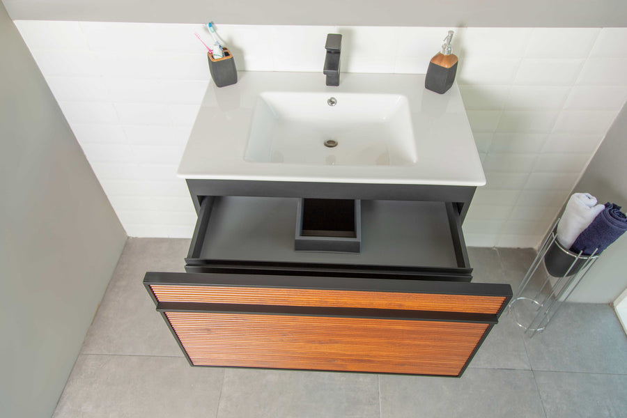 Modern bathroom sink with a white basin, black faucet, two toothbrush holders, and a wooden drawer partially open below, revealing organized compartments. Rolled towels are on a metal stand to the right.