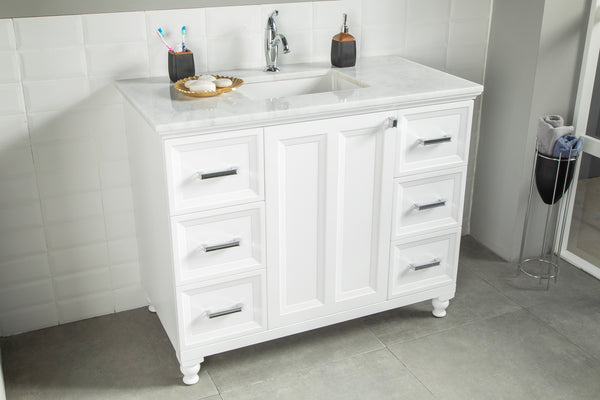 A white bathroom vanity with a marble countertop, chrome handles, built-in sink, soap dispenser, toothbrushes, and soap dish, standing on gray tile flooring against a white tiled wall.