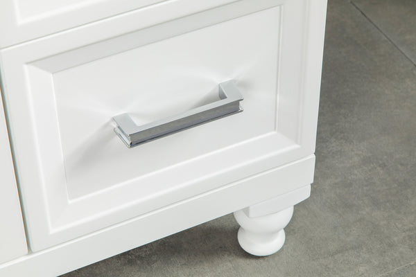 Close-up of a white drawer with a modern, rectangular silver handle. The drawer is part of a piece of furniture with paneled detailing and rounded feet, standing on a gray tiled floor.
