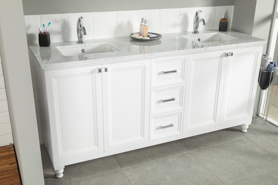 White double-sink bathroom vanity with marble countertop, chrome faucets, three central drawers, and cabinet doors. Toiletries, toothbrushes, and soap dispenser are placed on top. Grey tiled floor and white tiled backsplash.