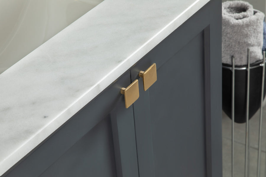 Close-up of a bathroom vanity with a white marble countertop, dark gray cabinet doors, and gold square handles. In the background, rolled towels are placed in a black metal rack.