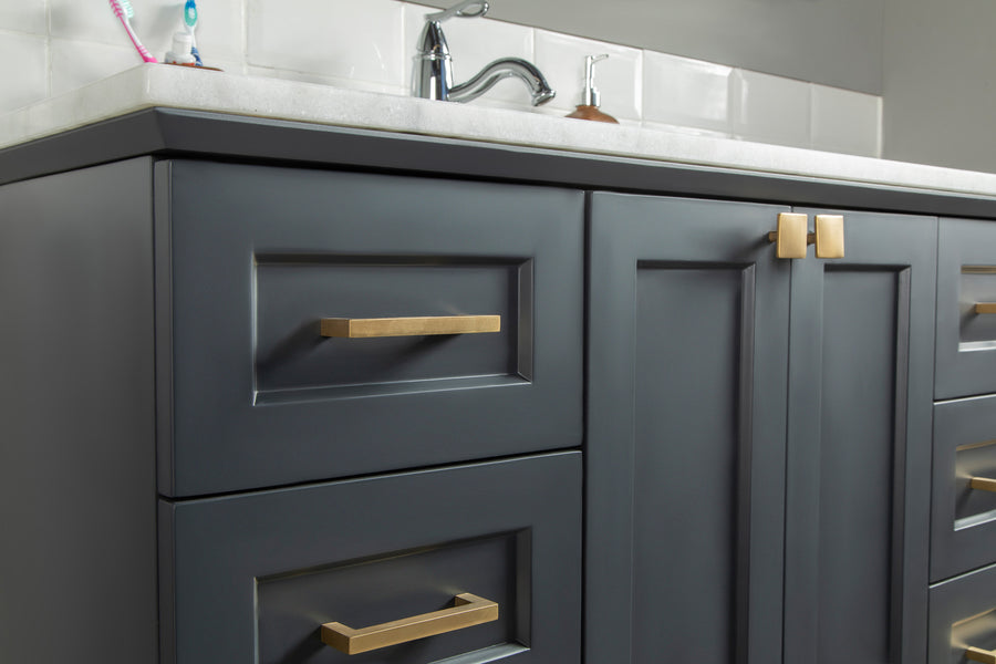 Close-up of a modern bathroom vanity with matte gray cabinets, gold handles, and a white countertop. A chrome faucet and toothbrush in a holder are visible on the counter in the background.