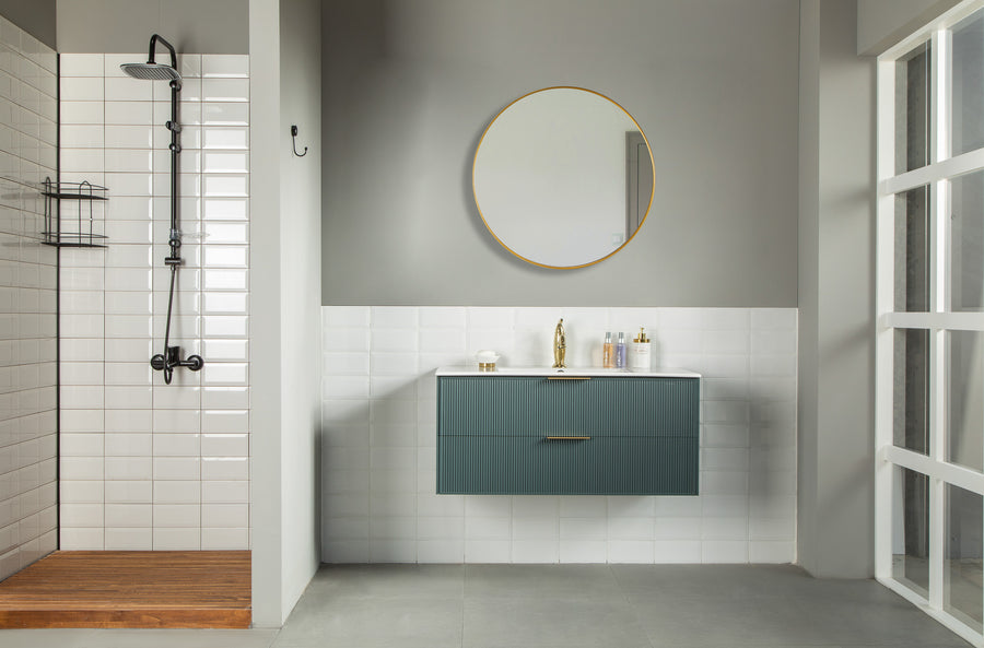 Modern bathroom with a walk-in shower on the left, a floating teal vanity with a gold faucet in the center under a round mirror, and a glass door on the right. The walls are gray and white tiles, and the floor is light gray.