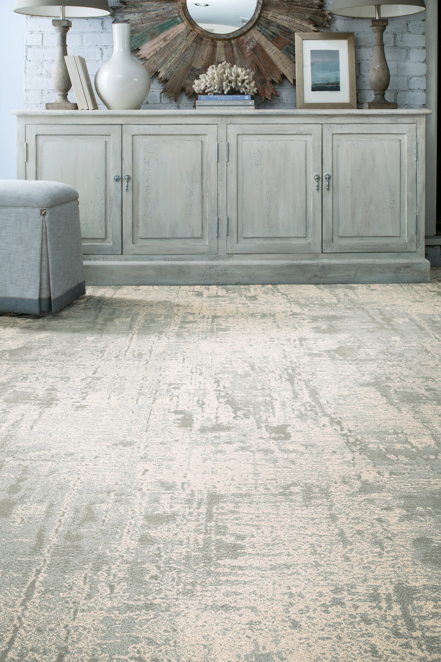 A light-colored, textured rug covers the floor in front of a distressed wooden sideboard, decorated with vases, a lamp, a small plant, and a round mirror. A gray upholstered bench sits to the left.