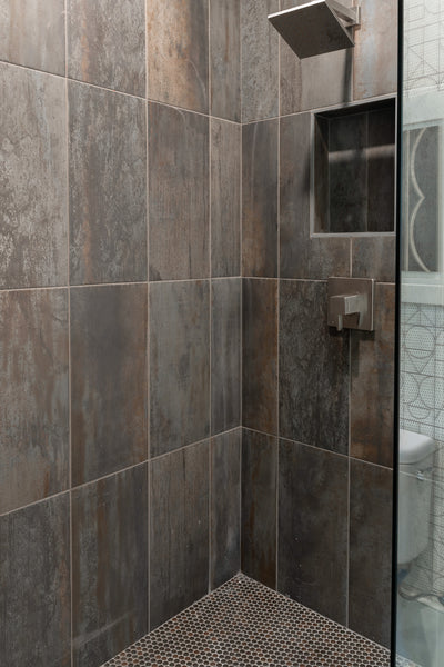 A modern shower with large dark, textured tiles, a square rainfall showerhead, built-in wall shelf, and geometric-patterned floor tiles. Part of a glass door and a glimpse of a toilet are visible on the right.