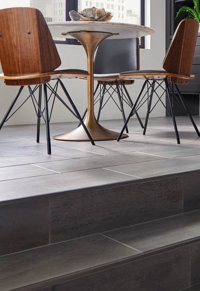 A modern dining area with a round marble-top table and three wooden chairs with black metal legs, set on gray tiled flooring near a window in a bright room.