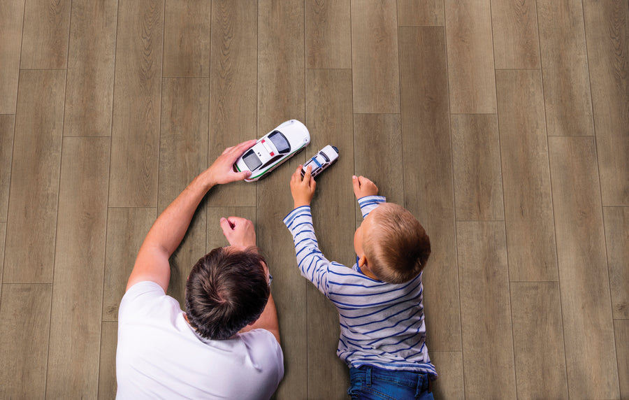A man and a young boy lie on a wooden floor, playing with toy police cars. The man holds a larger car while the boy holds a smaller one. Both are seen from above, enjoying their playtime together.