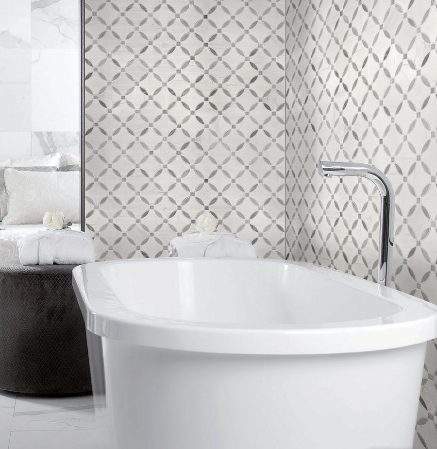 A modern bathroom with a white freestanding bathtub, geometric patterned gray and white wall tiles, a chrome faucet, and rolled white towels on a dark round table.