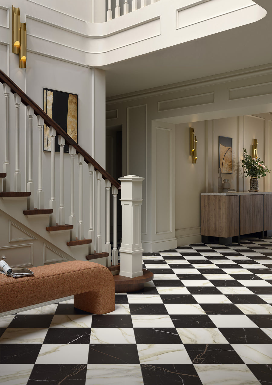 A modern entryway features a black and white checkered Palazzo 12x12 Glazed Porcelain Tile floor, seamless installation, a wooden staircase with white railings, rust-colored bench, console table with decor, and abstract art on cream walls.