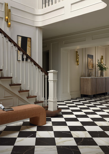 A stylish foyer features Palazzo 12x12 Glazed Porcelain Tile in a black and white checkered pattern, a curved wooden staircase with white railings, a rust-colored bench, modern wall art, and a wooden console table with a vase of flowers.