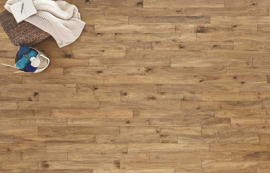 A basket with yarn, knitting needles, and a knitted blanket sits on the top left corner of a light brown wooden floor. The rest of the floor is clear and empty.