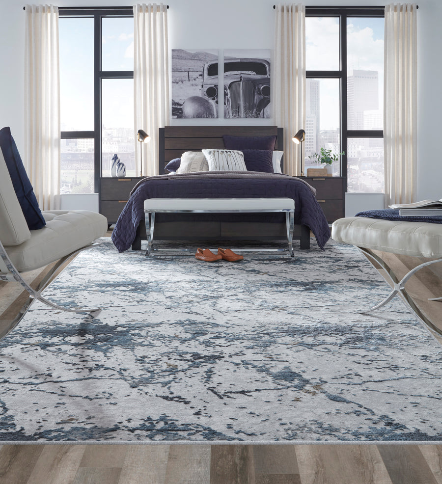 Modern bedroom with a large bed, blue and white bedding, a patterned rug, two white chairs, and large windows with city views. Shoes are placed at the foot of the bed, and artwork hangs above the headboard.