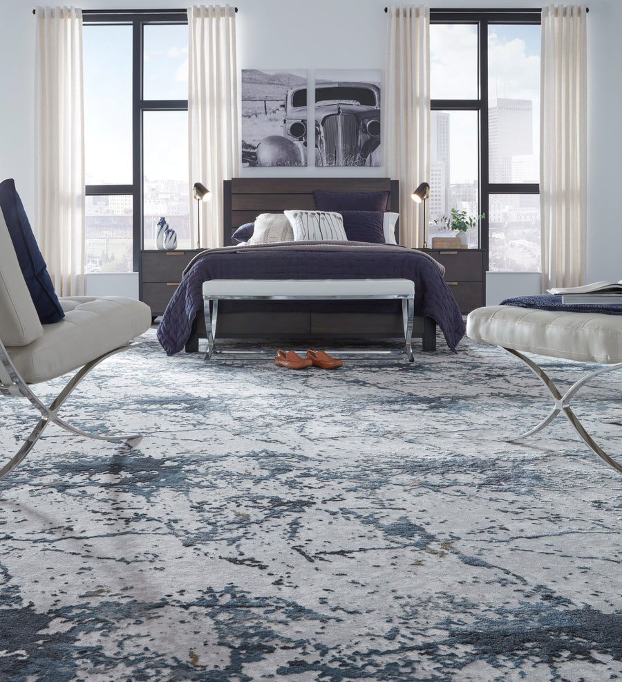 Contemporary bedroom with a large bed, blue and white bedding, two modern chairs, and cityscape artwork. Light curtains cover tall windows, and a pair of brown shoes is on the patterned carpet near the bed.