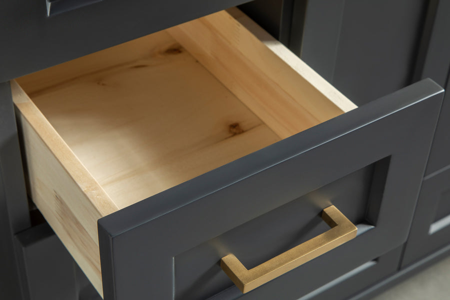 A close-up of a partially open drawer in a dark gray cabinet with a light wood interior and a rectangular gold handle.