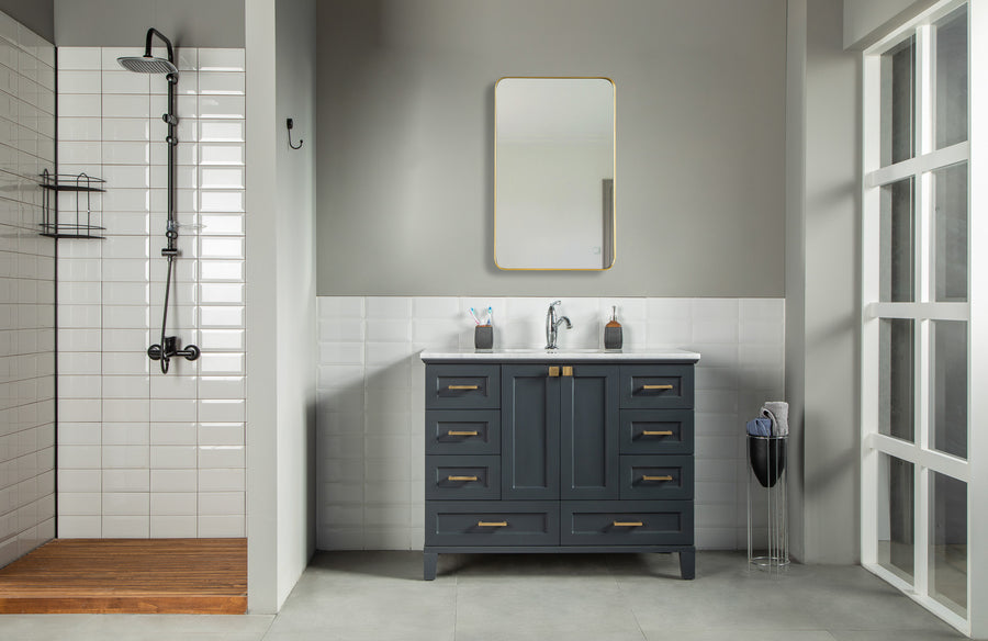 Modern bathroom with a glass shower on the left, classic white tiles, the Paloma 42" Vanity with soft-close drawers and gold handles, a rectangular mirror above, plus a trash bin and toilet brush on the right.