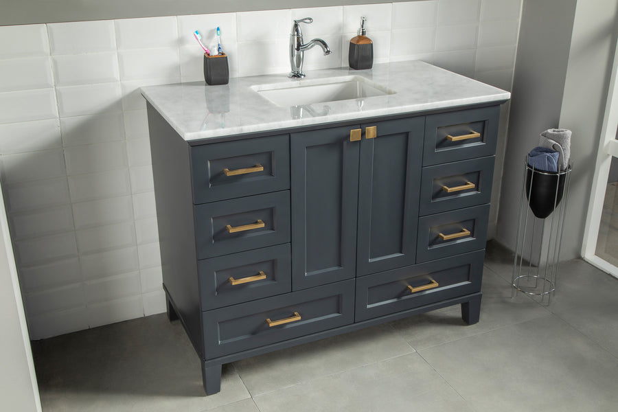 A modern bathroom vanity with a white marble countertop, rectangular sink, gray-blue cabinets with gold handles, a silver faucet, and two containers with toothbrushes and soap dispenser on top. A towel basket is nearby.
