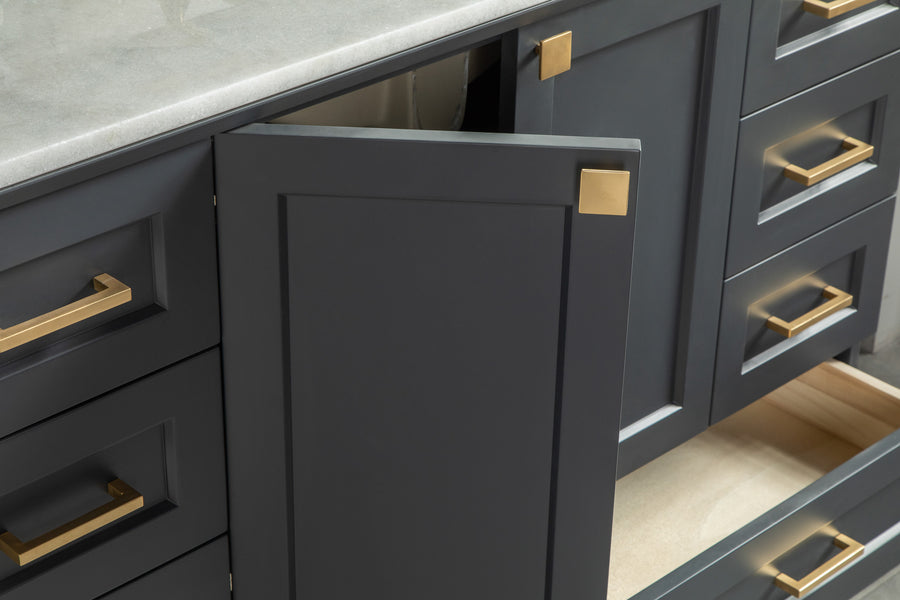 A close-up of dark gray kitchen cabinets with gold handles and knobs; one cabinet door is partly open and a bottom drawer is pulled out, showing the cabinet’s interior.