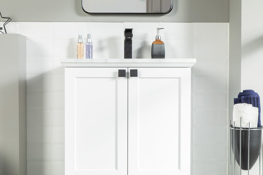 A modern white bathroom vanity with black handles, a white countertop, soap dispensers, and a sink faucet. Rolled towels are stacked in a metal holder to the right, and a mirror is partly visible above.