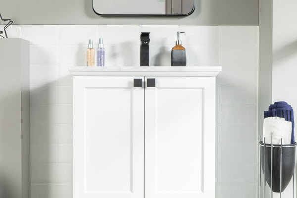 A modern white bathroom vanity with black handles, a white countertop, soap dispensers, and a sink faucet. Rolled towels are stacked in a metal holder to the right, and a mirror is partly visible above.