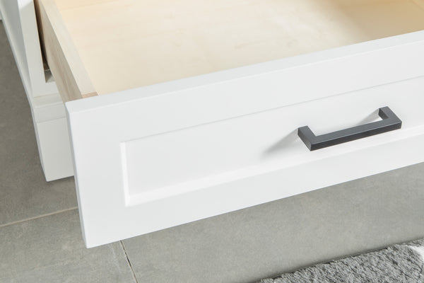 A close-up of an open white drawer with a black handle, showing its empty light wood interior. The drawer is set against a tiled floor with a gray textured mat nearby.
