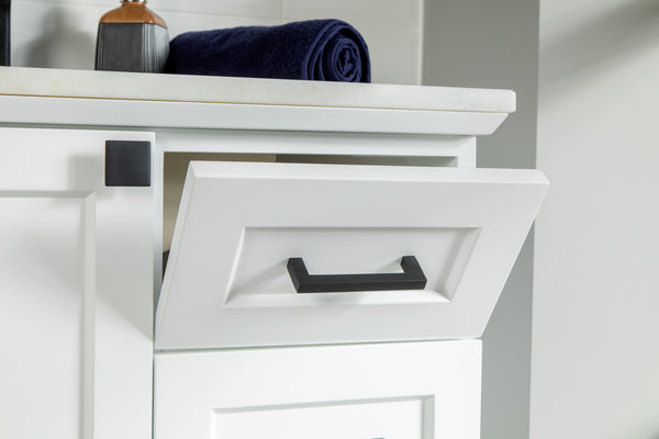 Close-up of a white bathroom or kitchen cabinet with a black handle; the top drawer is partially open and a dark blue towel is rolled up on the white countertop above.