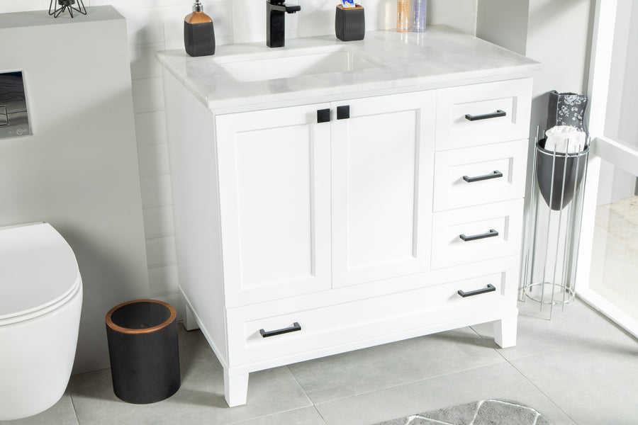 White bathroom vanity with black handles, a sink, and marble countertop next to a toilet. A brown wastebasket and a small towel stand are nearby on a gray tiled floor. Bottles and soap dispenser sit on the counter.