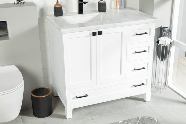 White bathroom vanity with black handles, a sink, and marble countertop next to a toilet. A brown wastebasket and a small towel stand are nearby on a gray tiled floor. Bottles and soap dispenser sit on the counter.