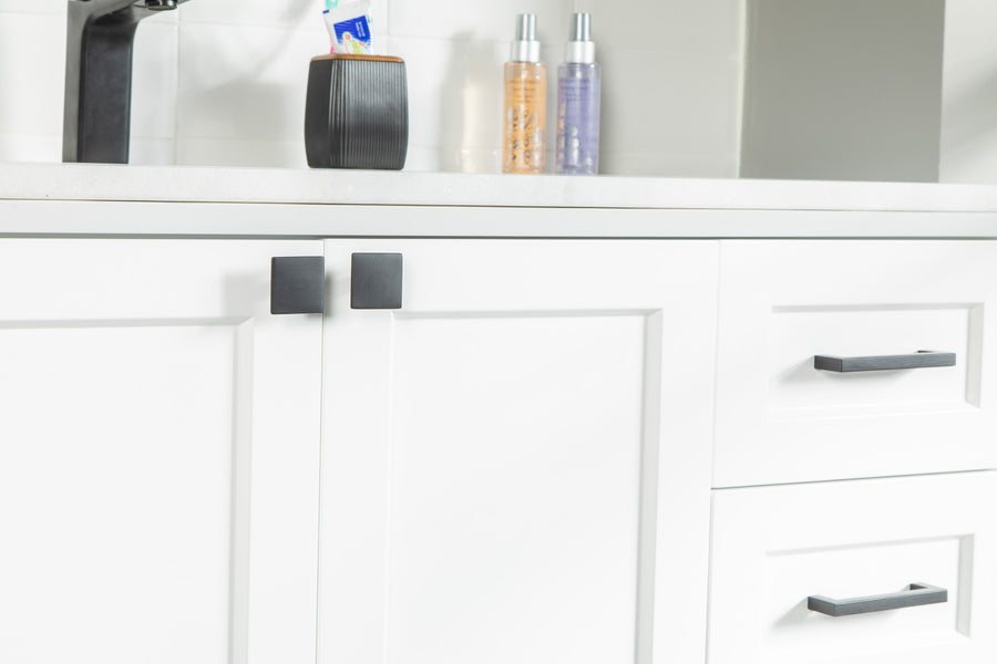Close-up of white bathroom cabinets with black handles and square knobs, a white countertop above, and soap dispensers and a black cup in the background.