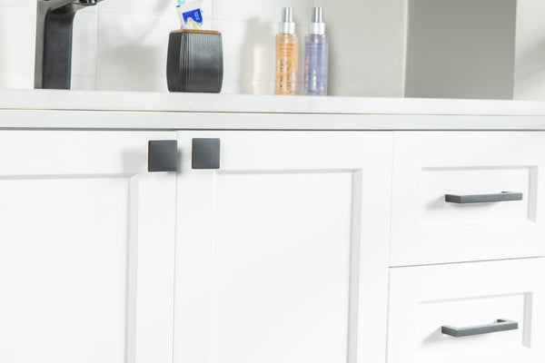 Close-up of white bathroom cabinets with black handles and square knobs, a white countertop above, and soap dispensers and a black cup in the background.
