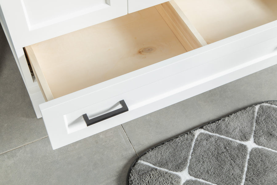 An open white drawer with a black handle is shown above a gray tiled floor next to a gray and white patterned rug. The drawer is empty and part of a white cabinet.