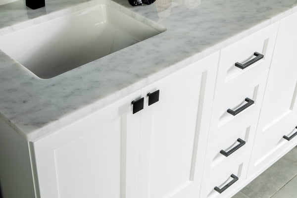 Close-up of a white bathroom vanity with a marble countertop, black square cabinet handles, and drawer pulls, featuring an integrated rectangular sink.