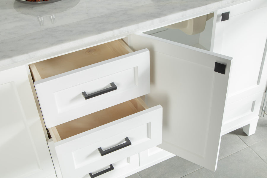 White kitchen cabinets with black handles are shown; two drawers and one door are open, revealing empty wooden interiors. The cabinets are topped with a marble countertop and set on a tiled floor.