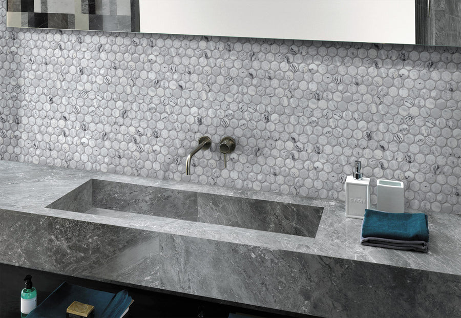 Modern bathroom sink with a rectangular stone basin, wall-mounted faucet, round marble tile backsplash, and countertop items including soap dispensers and neatly folded towels.