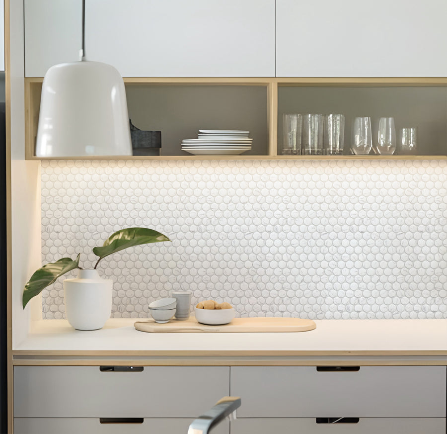 A modern kitchen with white cabinets, a round white pendant light, a white backsplash with circular tiles, a potted plant, glassware, plates, and small bowls arranged neatly on the counter and shelves.