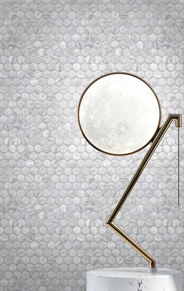 A modern, gold and black metal lamp with an angled arm and round white lampshade stands on a white marble surface, set against a gray marble tile wall with a circular pebble pattern.