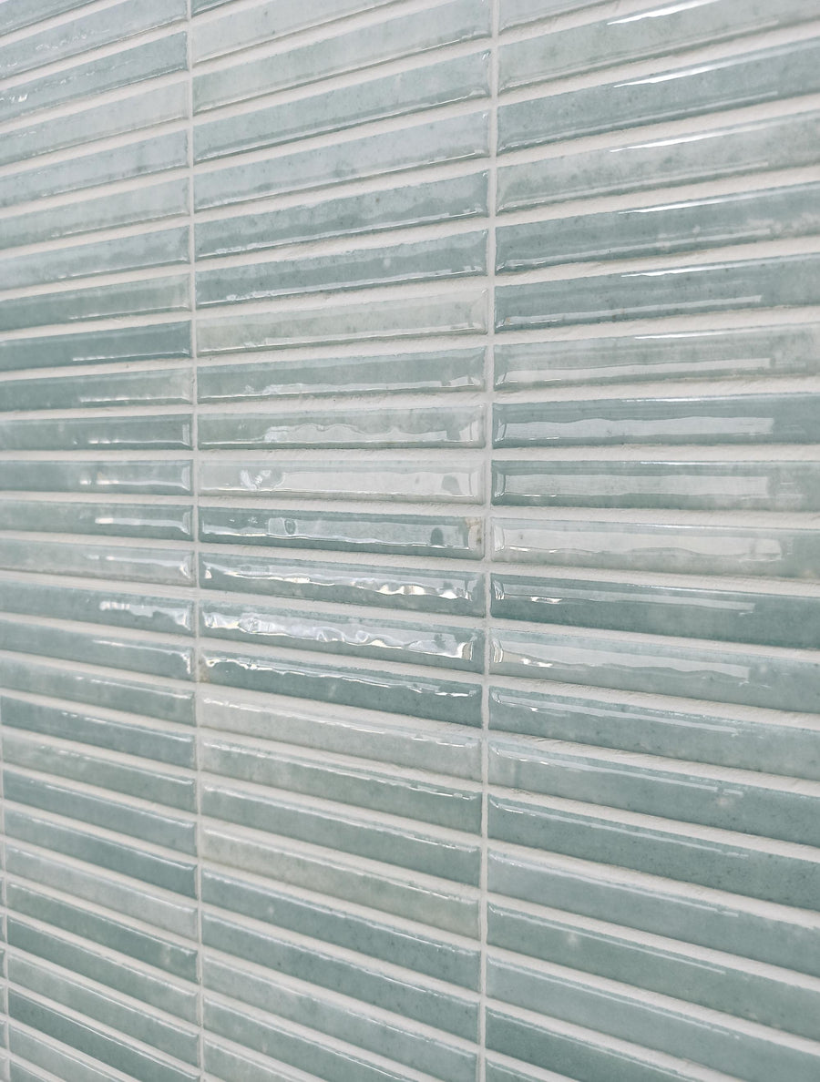 Close-up of a wall covered with glossy, rectangular ceramic tiles in shades of light teal and white, arranged in a horizontal pattern with visible grout lines.
