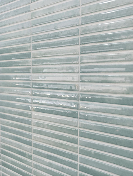 Close-up of a wall covered with glossy, rectangular ceramic tiles in shades of light teal and white, arranged in a horizontal pattern with visible grout lines.