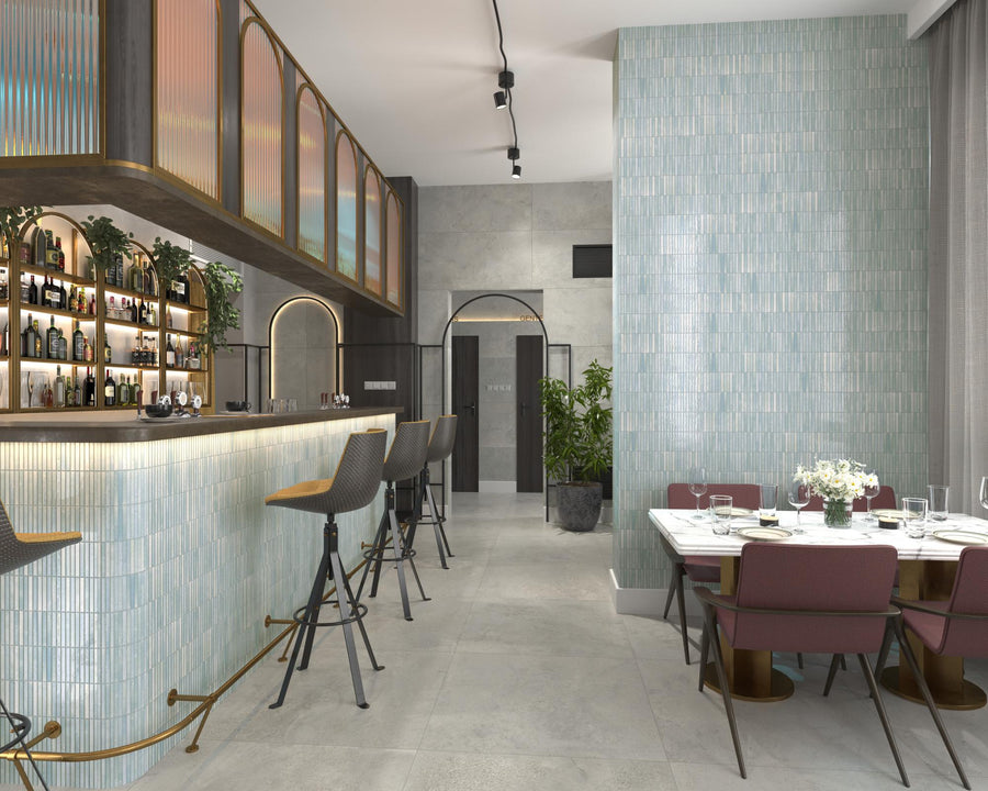 Modern restaurant interior with a light blue tiled bar, high chairs, a backlit shelf with bottles, potted plants, and a dining table set with pink chairs and a flower centerpiece.