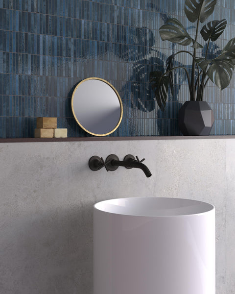 A modern bathroom with a white cylindrical sink, wall-mounted black faucet, round mirror with a gold rim, and a geometric black vase with green leaves against blue tiled and light stone walls.