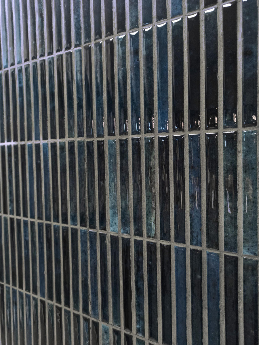 Close-up of a wall covered with rectangular, glossy, dark blue ceramic tiles arranged in a vertical grid pattern. The tiles have subtle variations in shade and reflect light, creating a textured appearance.
