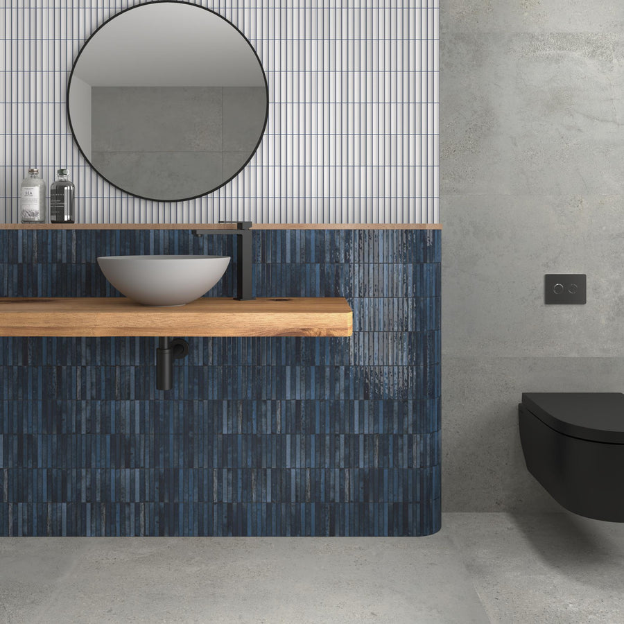Modern bathroom with a round mirror above a wooden countertop, vessel sink, and matte black faucet. The wall has blue tiles below and vertical white tiles above. A black wall-mounted toilet is on the right.