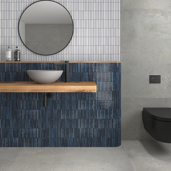 Modern bathroom with a round mirror above a wooden countertop, vessel sink, and matte black faucet. The wall has blue tiles below and vertical white tiles above. A black wall-mounted toilet is on the right.