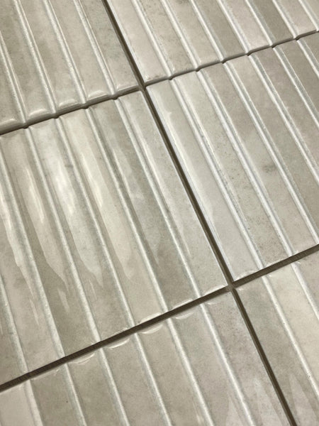 Close-up view of light gray rectangular tiles with a ribbed texture, arranged in a grid pattern. The tiles have a glossy finish and visible grout lines between them.