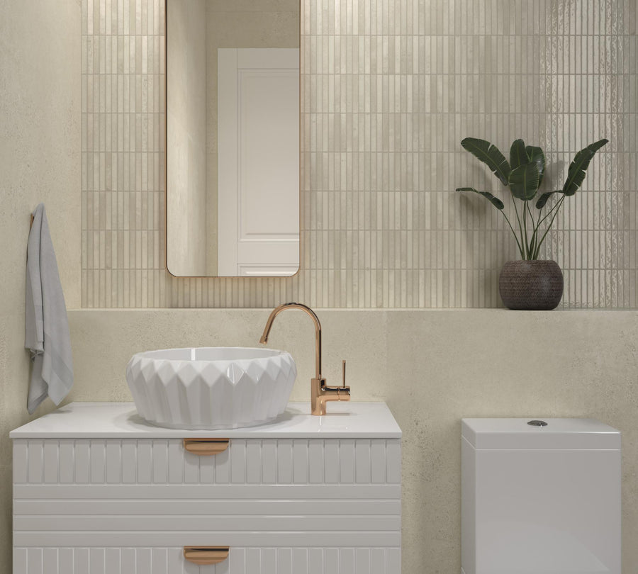 A modern bathroom with a white geometric sink, rose gold faucet, rectangular mirror, potted plant, wall towel, and a feature wall of Dunes 12x24 Porcelain Tile in textured beige.