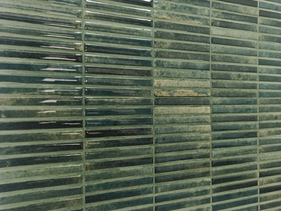 Close-up of a wall covered in glossy, rectangular, green ceramic tiles arranged in a horizontal, staggered pattern. The tiles have varied green tones and a textured, slightly uneven surface.