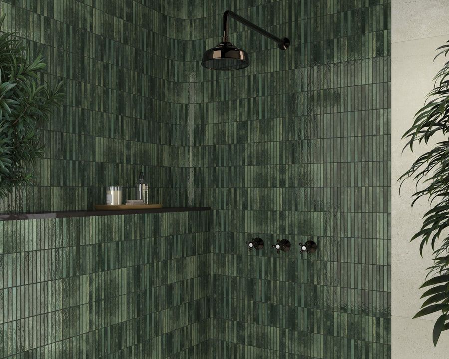 A modern shower with glossy green rectangular tiles, a black rainfall showerhead, and a built-in shelf holding toiletries. Lush green plants are positioned on both sides of the shower area.