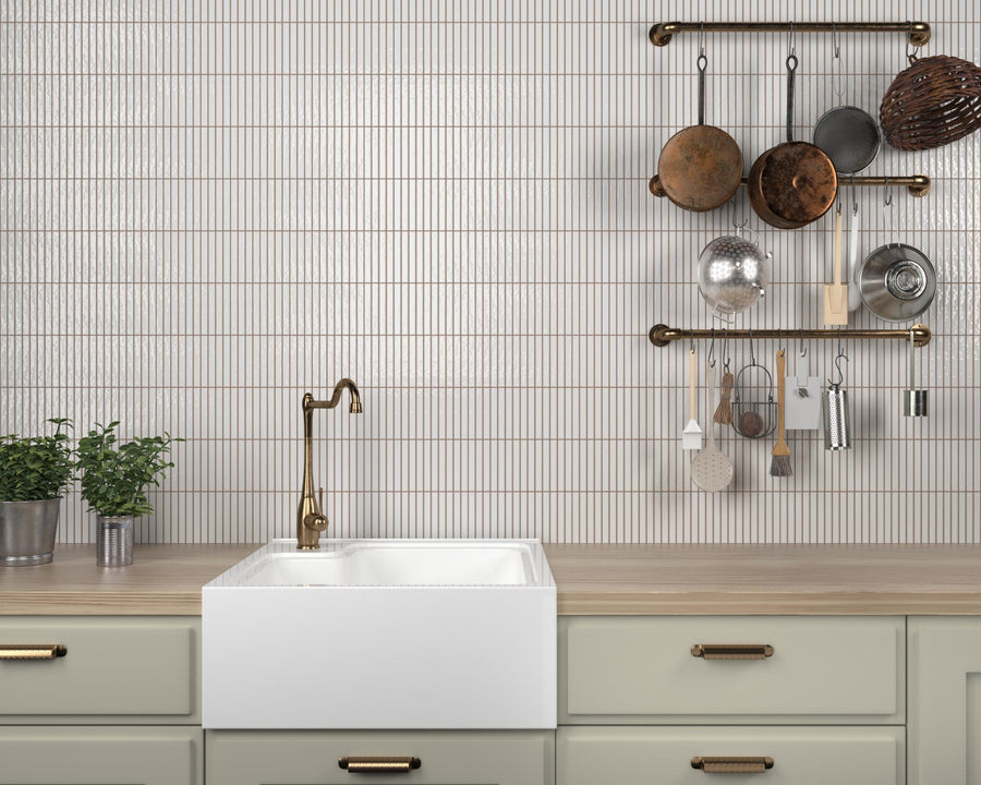 A modern kitchen features a farmhouse sink, brass faucet, light wood countertop, potted plants, and a wall with vertical white tiles, shelves holding various hanging pots, pans, and utensils.