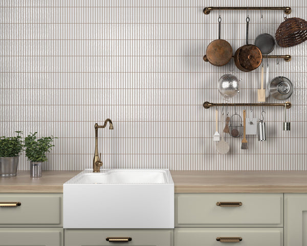 A modern kitchen features a farmhouse sink, brass faucet, light wood countertop, potted plants, and a wall with vertical white tiles, shelves holding various hanging pots, pans, and utensils.