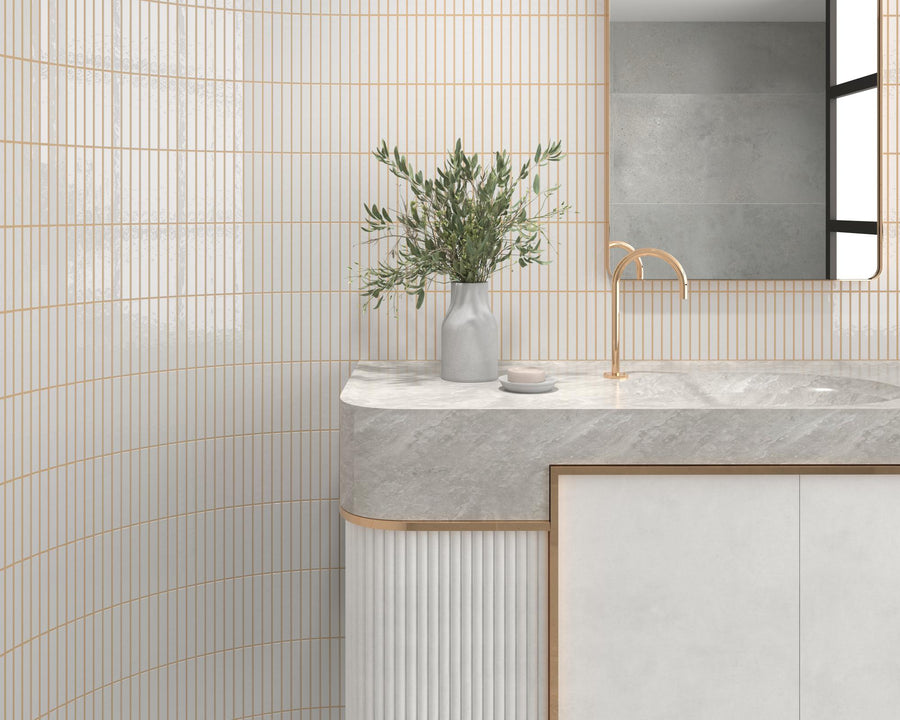 A modern bathroom with vertical white and gold tiles, a marble countertop, a gold faucet, a rectangular mirror, and a vase with green branches on the counter.